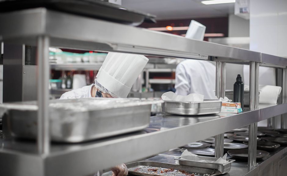 Interior view of a commercial kitchen corner with white subway tiles on the walls and patterned floor tiles. A person is partially visible behind a door, using a mop to clean the tiled floor. The kitc