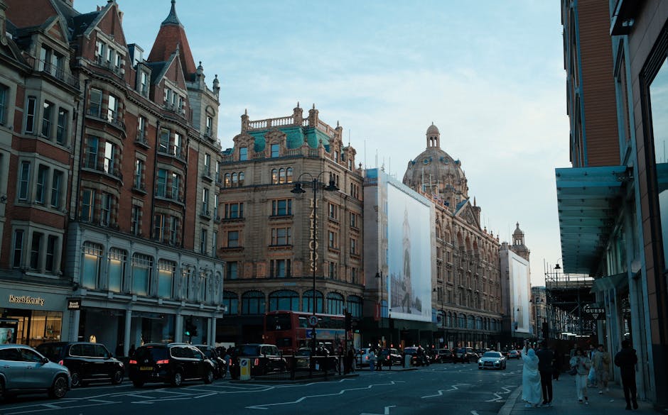 A busy street view in Knightsbridge featuring historic and modern architecture. Classic stone buildings with decorative facades and large windows line the street, with some featuring ornate towers and domed roofs. The scene shows pedestrians walking along the pavement, and a variety of vehicles, including cars and a double-decker bus, are present on the road. The lighting suggests late afternoon with a partly cloudy sky, creating a balanced natural light that highlights the building details. On the right side of the image, a contemporary glass storefront with a blue awning is visible, contrasting with the traditional architecture. The overall cleanliness of the street and sidewalks emphasizes a well-maintained urban area, consistent with the refined atmosphere of Knightsbridge. This image illustrates the setting where professional domestic and commercial cleaning services, such as those offered by Oven Cleaning Knightsbridge, are essential for maintaining hygiene and aesthetic appeal.