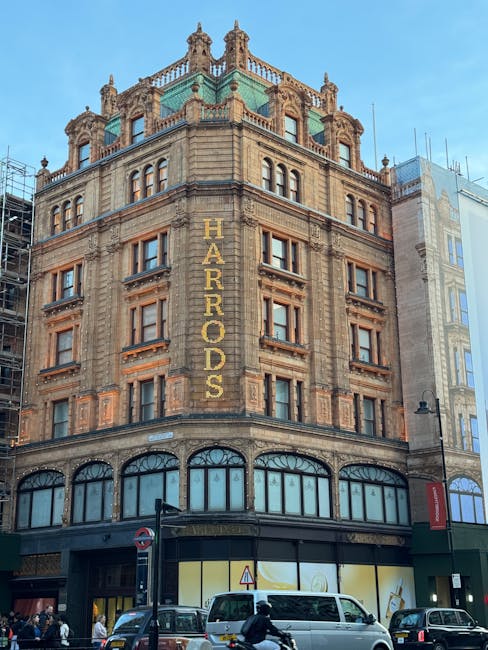 Exterior view of the historic Harrods department store building in Knightsbridge, featuring ornate stonework, large arched windows on the ground floor, and decorative architectural details at the upper levels. The signage displaying 'HARRORDS' is prominently visible on the corner of the building. The scene includes vehicles and pedestrians on the street, with a sunny sky overhead. This image exemplifies the classic architecture of Knightsbridge, aligning with the professional cleaning and maintenance focus of Oven Cleaning Knightsbridge at [PAGE_URL].